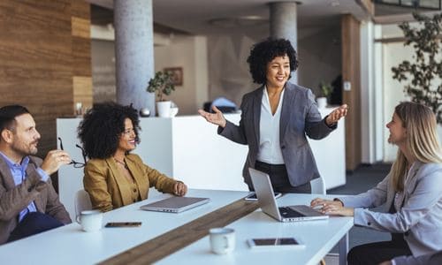 A confident businesswoman leads a coaching conversation with three colleagues seated around a table, gesturing openly as she speaks.