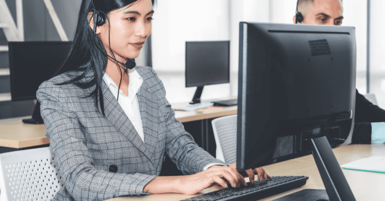 Young salesperson wearing a headset sitting at her desk making cold calls and typing notes on the computer in an office
