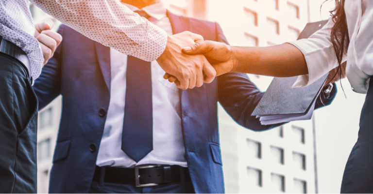 Three salespeople outside standing in front of an office building in suits shaking hands one is holding a clipboard