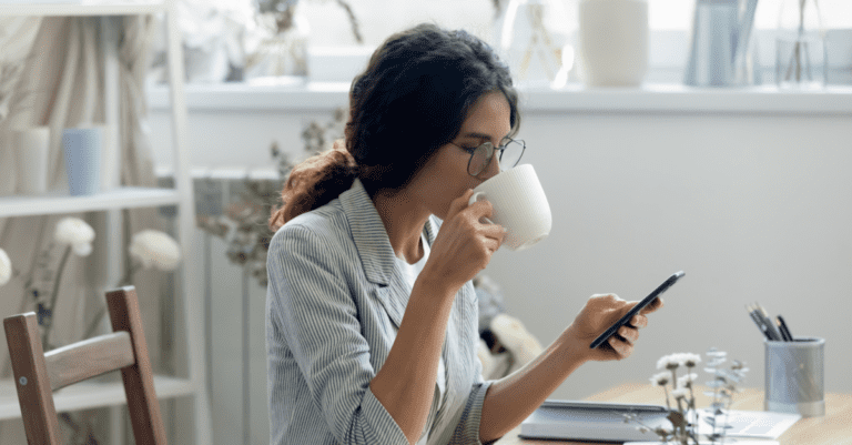 Young sales person sitting at desk wearing a gray and white stripped jacket drinking coffee and texting prospects
