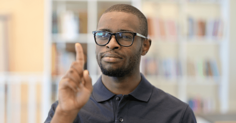 Young man with a black golf shirt and black glasses shaking finger no in an office in front of an out of focus bookshelf