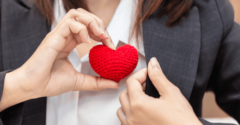 Business woman with a gray suit and white shirt holding a heart in her right hand while holding the left side of her jacket