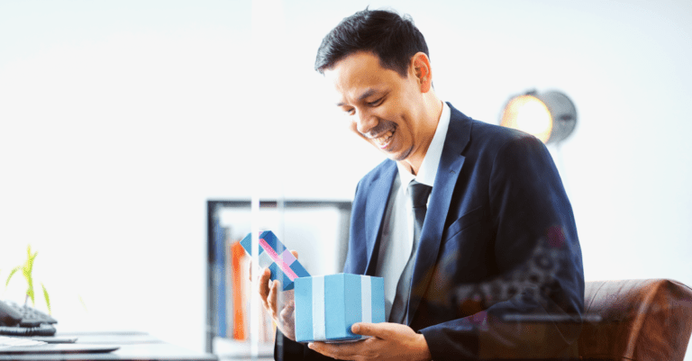 Male businessman wearing a blue suit opening a light blue holiday gift with white ribbon sitting at his desk in his office